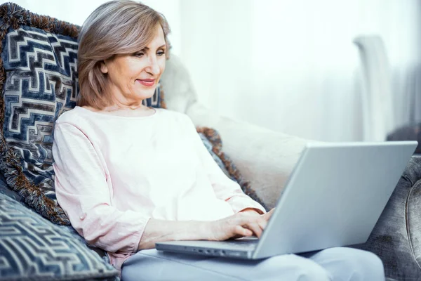 Una mujer mayor de 50 años se sienta en una computadora portátil en casa en la sala de estar . — Foto de Stock