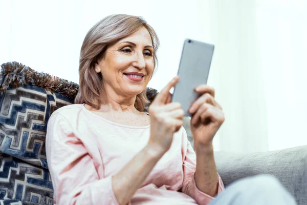 Mujer mayor con un teléfono en sus manos en buen estado de ánimo está descansando en casa en la sala de estar . — Foto de Stock