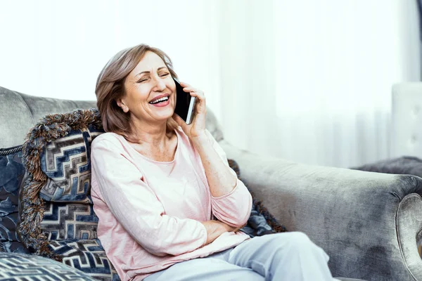 Mulher idosa com um telefone nas mãos de bom humor está descansando em casa na sala de estar . — Fotografia de Stock