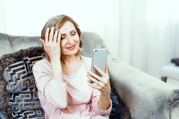 Mulher idosa com um telefone nas mãos de bom humor está descansando em casa na sala de estar . — Fotografia de Stock