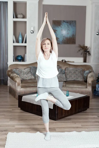 O femeie de 50 de ani practică yoga acasă în sufragerie . — Fotografie, imagine de stoc