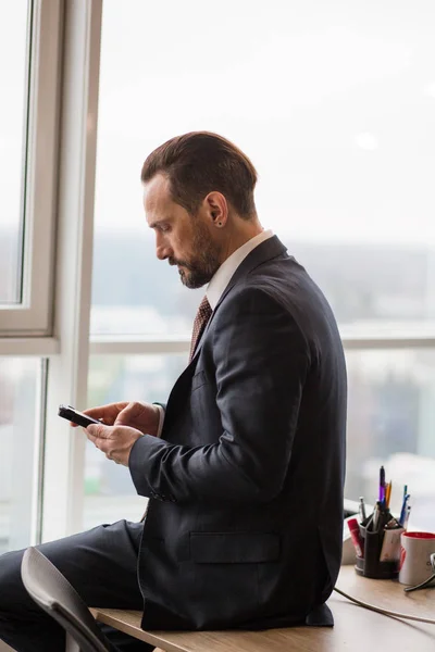 Een goed geklede zakenman surft op zijn mobiele telefoon op het moderne kantoor, achter is een raam, hij is gefocust en serieus. — Stockfoto