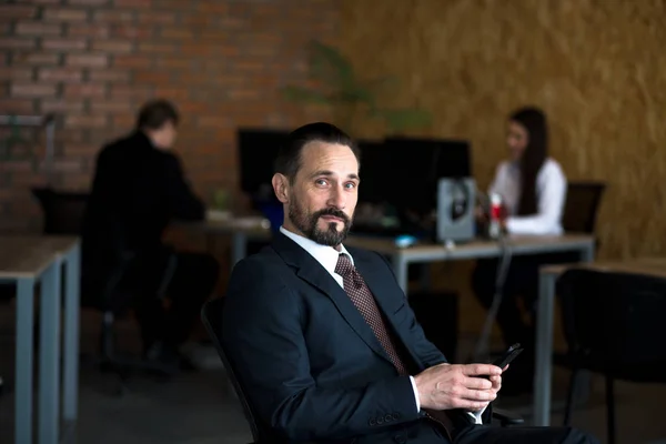 Handsome businessman sitting on his chair in front of his office. Two colleagues are working behind him. — Stock Photo, Image