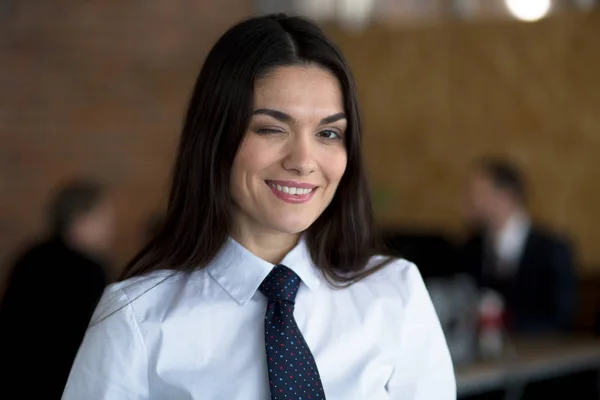 Alegre mujer de negocios sonriente guiñando un ojo en una blusa blanca y una corbata negra. Dos colegas están trabajando detrás de ella. . — Foto de Stock