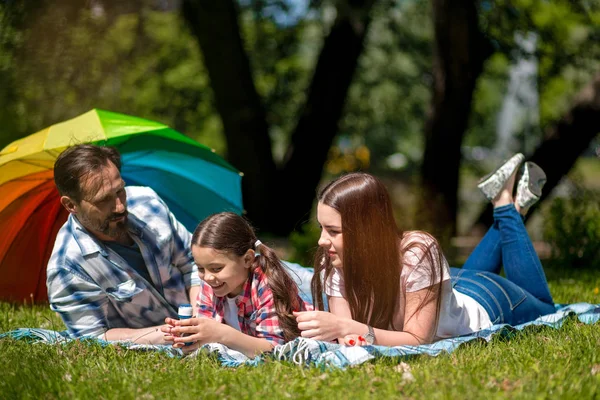 Familj som ligger tillsammans på en picknick filt utomhus i parken. Färgglada paraply på bakgrunden. — Stockfoto
