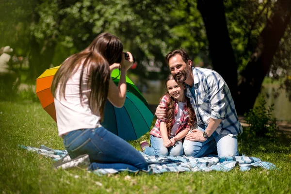 Joven madre haciendo foto de marido e hija en el parque. Padre está abrazando a su hija mientras está sentado en la manta. Paraguas colorido está junto a ellos . — Foto de Stock