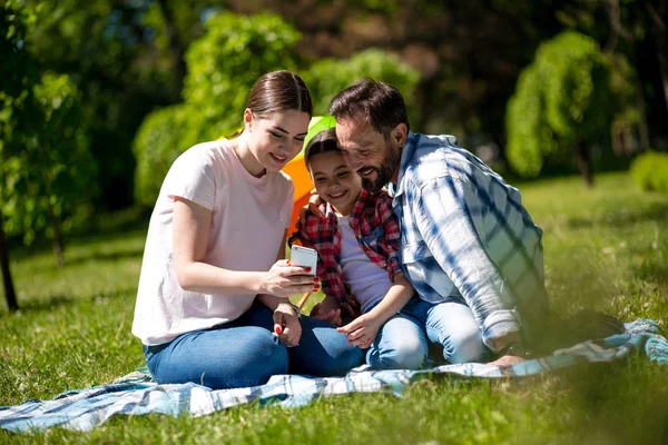 Ung Kvinna Visar Foton På Telefon Till Hennes Make Och Dotter Medan Sitter På Filten I Parken. — Stockfoto