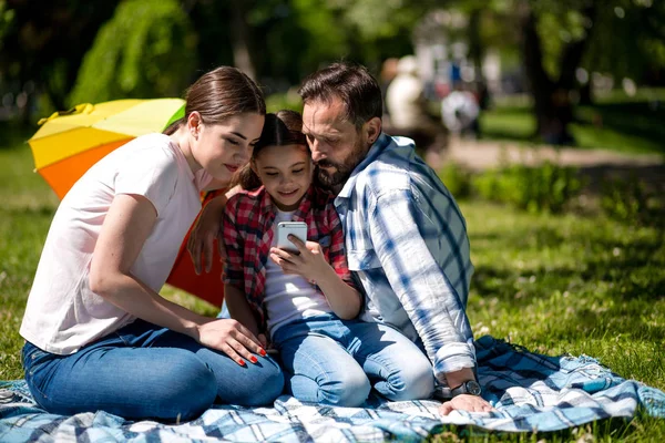 Leende familj tittar på foto på mobiltelefon medan du sitter på täcket i parken. — Stockfoto