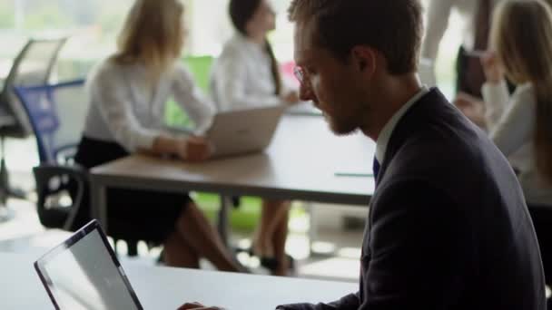 En affärsman som skriver på sin laptop. Chefen förklarar ny idé för arbetstagare i bakgrunden. — Stockvideo