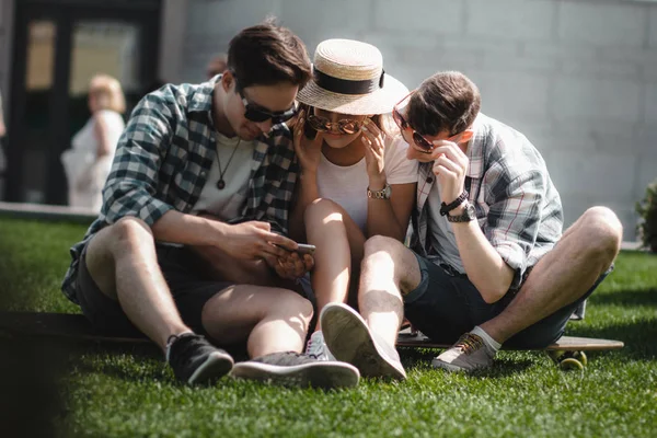 Три молоді друзі сидять на траві на відкритому повітрі і дивляться на мобільний телефон . — стокове фото
