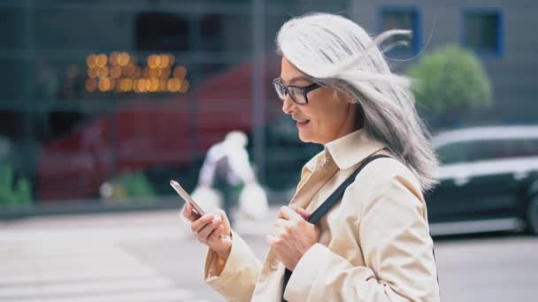 La mujer asiática de mediana edad está de pie en la calle y escribiendo en su teléfono inteligente — Vídeos de Stock