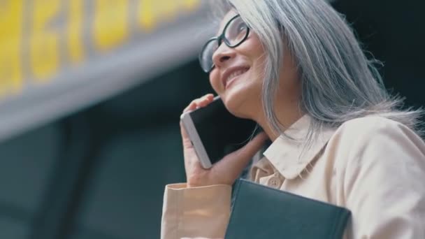 Die asiatische Frau mittleren Alters hält ein Notizbuch in der Hand und telefoniert — Stockvideo