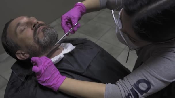 Il parrucchiere taglia la barba al cliente tagliando o raddrizzando il collo. Un uomo visita un salone di bellezza durante un periodo di quarantena pandemica. Prore 422 — Video Stock