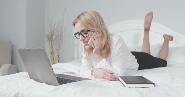 Donna è sdraiata su un letto con un computer portatile e parlare al telefono. La donna riposa in un hotel dopo un lungo volo prendendo un appuntamento con i clienti. Prore 422 — Video Stock