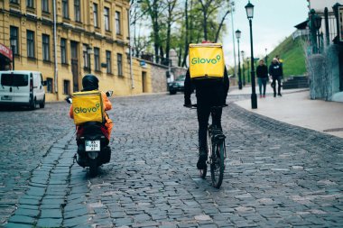Glovo courier service. Two food deliverymen with Yellow backpacks on a motorbike on a bicycle ride along a city street. Rear view. Kyiv, Ukraine, 24th of April 2020, Andreevskiy spusk Street clipart