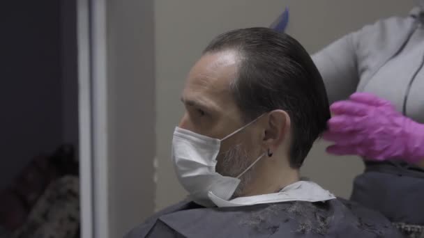 Πελάτης σε μια προστατευτική μάσκα κάθεται υπομονετικά, ενώ κομμωτήριο βούρτσισμα των μαλλιών από στυλίστα και για να ετοιμαστείτε να κοπεί. Ο άνθρωπος επισκέπτεται ινστιτούτο ομορφιάς κατά τη διάρκεια περιόδου πανδημίας καραντίνας. Πρότυπα 422 — Αρχείο Βίντεο