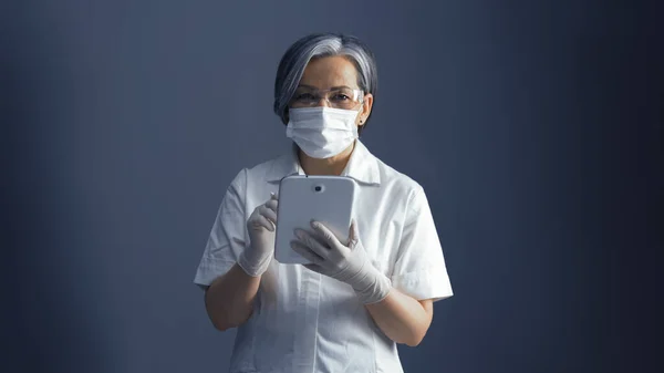 Médico feminino trabalha com bloco de notas eletrônico. Mulher bonita em uniforme branco usando aplicativos em gadget digital inteligente em seu trabalho de pé sobre fundo cinza azul com espaço de cópia em ambos os lados. Imagem tonificada — Fotografia de Stock