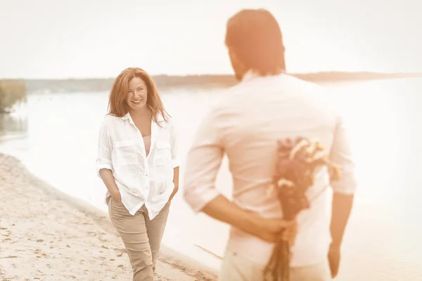 Romantisk møte mellom et kjærlig par på en sandstrand ved havet. Fokuser på den modne, smilende kvinnen som ser på mannen med bukett med villblomster bak ryggen mens han står i forgrunnen. Tekstet bilde – stockfoto