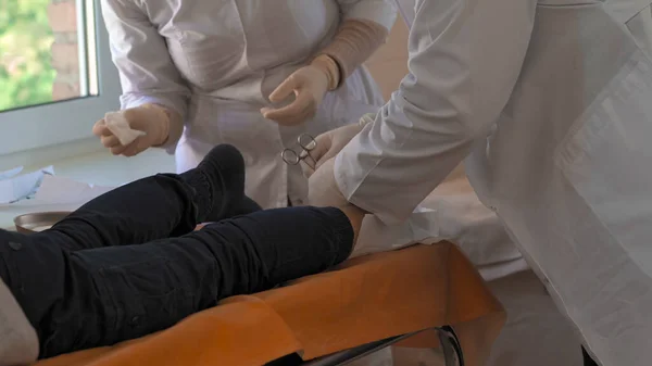 Doctor and nurse in white coats and disposable gloves perform medical manipulations with the patients leg. Surgeons office. Close up shot. City Hospital. May, 2020, Brovary, Ukraine — Stock Photo, Image