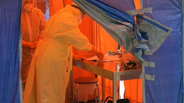 Médico en traje epidemiológico escribiendo datos para registrar. Imagen abstracta desenfocada. Corredor rojo en la entrada al hospital. Centro de contracción covid-19. Hospital Municipal. mayo, 2020, Brovary, Ucrania —  Fotos de Stock