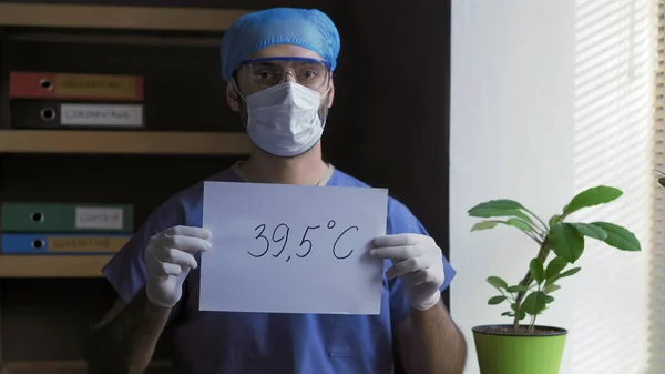Orvos figyelmeztet a magas láz tünete lehet a covid-19 veszélyes betegség. Orvosi dolgozó védő egyenruhában és maszkban egy papírlapot tart 39,5 fokos felirattal. — Stock Fotó