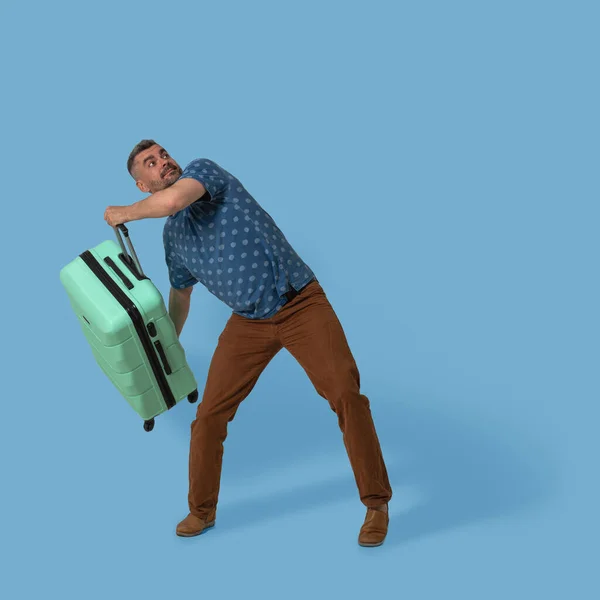 Man håller plast resväska poserar i studio på blå bakgrund. Vit man ska kasta sin gröna resväska med bagage på övervåningen. Resor reklam mall med textutrymme — Stockfoto