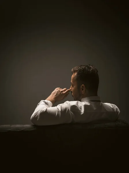 Empresário confuso sentado no sofá toca cara com mão pensativa. Visão traseira do homem caucasiano preocupado em camisa branca sentado no sofá no quarto escuro. Imagem tonificada — Fotografia de Stock