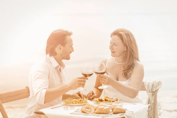 Очаровательная пара соединила бокалы красного вина, глядя друг на друга. Счастливый зрелый мужчина и женщина вместе имеют свидание в пляжном кафе у воды. Романтическая концепция. Тонированное изображение — стоковое фото