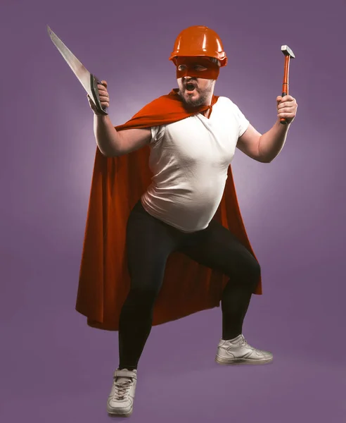 Super hero man holding construction tools. Engineer or Repairman in red helmet and super hero uniform with saw and hammer standing isolated on grape purple background