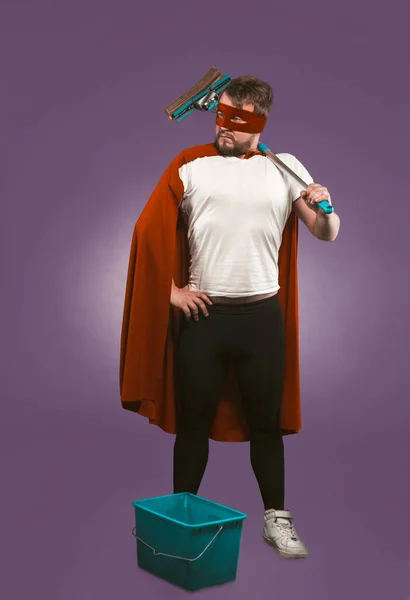 Super held reiniger professioneel. Vertrouwen man in rode superheld kostuum met emmer en een dweil staat op druivenpaarse achtergrond. Super reinigingsconcept — Stockfoto