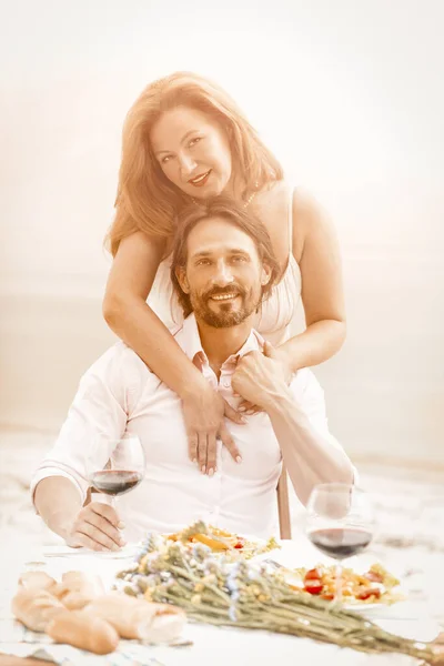 Pareja feliz abrazándose en la cafetería de la playa mientras mira la cámara. Precioso hombre y mujer abrazándose a la mesa con comida y copas de vino tinto sobre ella sobre fondo de mar o océano. Imagen tonificada — Foto de Stock