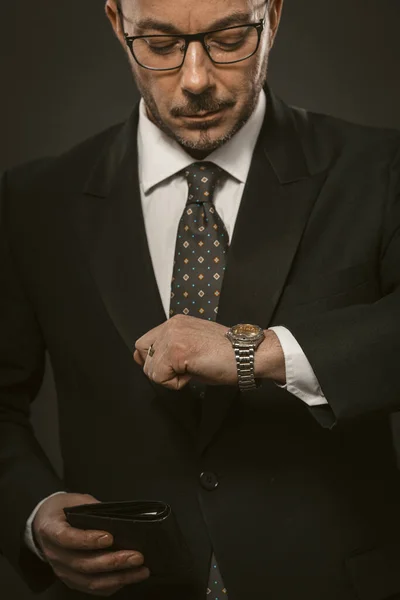Пунктуальний бізнесмен перевіряє час, дивлячись на наручний годинник, тримаючи гаманець в іншій руці. Добре одягнений бородатий чоловік в окулярах дивиться на ручний годинник. Крупним планом. Очікування на концепцію. Тоноване зображення — стокове фото