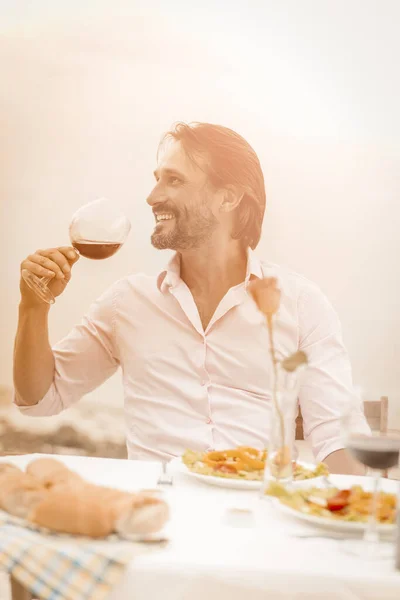 Den leende kaukasiske mannen håller ett glas rött vin vid bordet vid kusten utomhus och tittar åt sidan. Romantiskt dejtkoncept. Firandet av rostat bröd. Tonad bild — Stockfoto