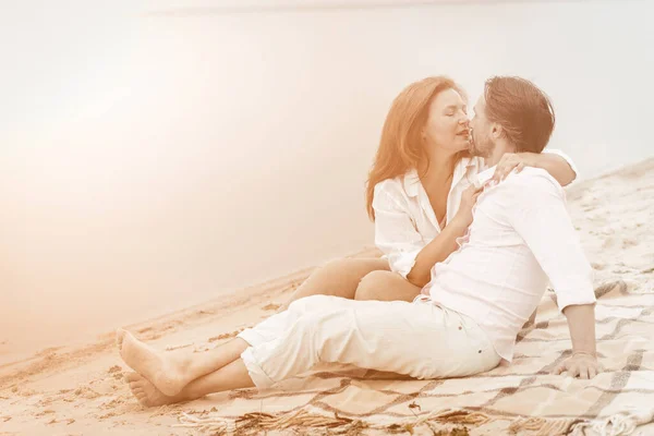 Couple in love gently hugs and kisses sitting on blanket in the sand near water. Mature couple in white in white clothes spend time together relaxing outdoors. Romantic date concept. Toned image — Stock Photo, Image