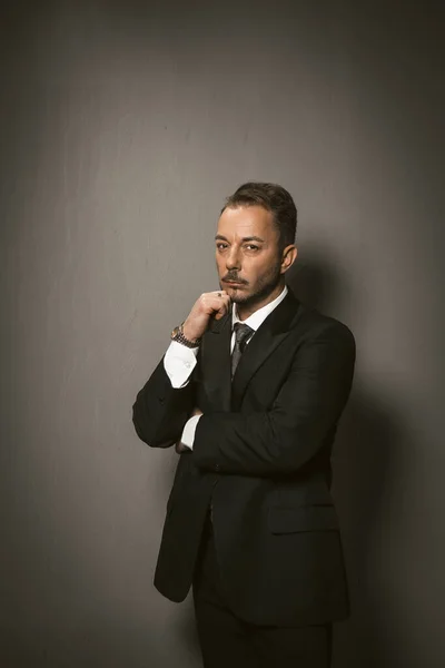 Sad businessman looks at camera feeling stress in crisis period. Serious caucasian man in black suit touches his chin with hand standing on grey background. Toned image — Stock Photo, Image