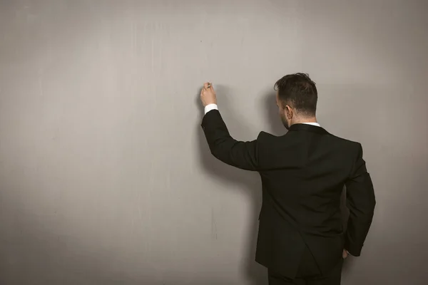 Achteraanzicht van een zakenman die op de muur schrijft. Creatieve man in zwarte elegantie pak tekening door pan of krijt op lichtgrijze muur. Template concept. Getinte afbeelding — Stockfoto