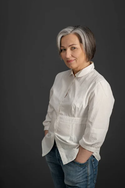 Mujer madura de pelo gris sonríe cogida de la mano en los bolsillos mientras mira a la cámara. Encantadora mujer caucásica en camisa blanca posando sobre un fondo gris —  Fotos de Stock