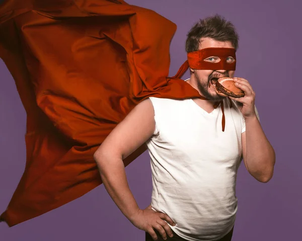 Homem super-herói faminto come hambúrguer grande com carne. Homem de manto voador vermelho come olhando para a câmera no fundo roxo de uva. Conceito de lanche fast food — Fotografia de Stock