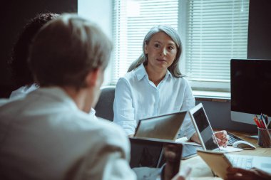 Office staff brainstorming. Elderly Asian woman listens attentively looking at her colleagues while sitting at negotiating table during a business meeting clipart