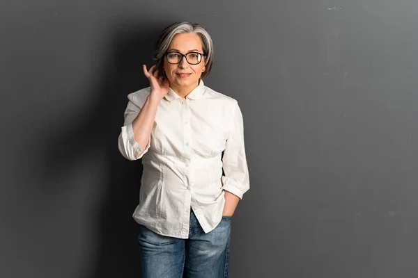 Mujer bastante madura corrige coqueta peinado de longitud media. Adorable modelo caucásico de pelo gris en jeans posando sobre fondo gris. Copiar espacio o espacio de texto en el lado derecho —  Fotos de Stock