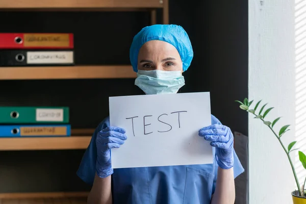 Médico en uniforme azul anuncia la necesidad de hacer pruebas médicas para identificar una enfermedad peligrosa en el contexto de la oficina médica. Doctora con inscripción TEST en papel en blanco. Concepto pandémico —  Fotos de Stock