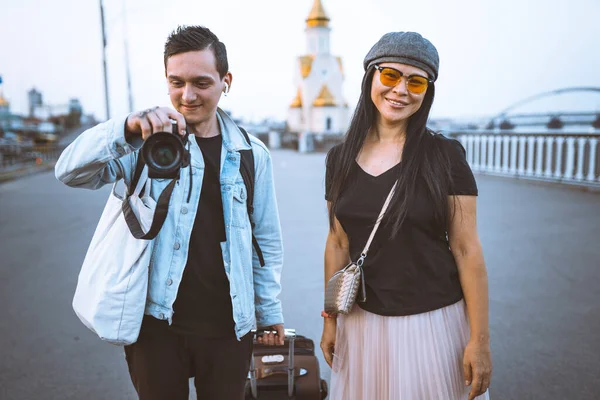 Nehir kenarında bavullu mutlu turistler. Genç adam fotoğraf çekiyor. Sıradan kıyafetler giyen bir kadın kameraya bakıp gülümsüyor. Seyahat kavramı — Stok fotoğraf