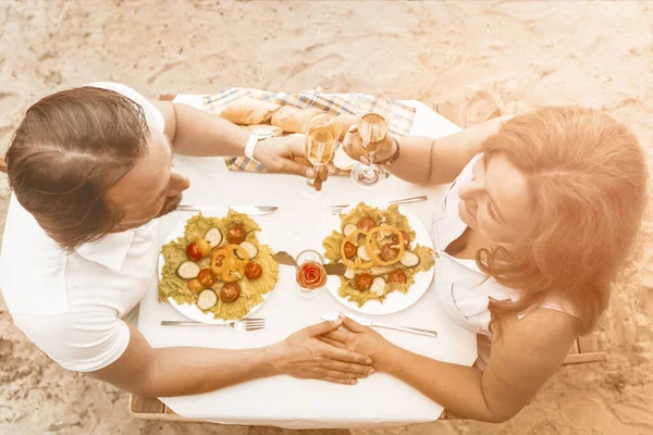 愛する成熟したカップルは、砂浜の屋外のカフェでロマンチックなデートをしています。男と女は砂のビーチでおいしい食べ物や飲み物とテーブルに座って手を握っています。角度の高いビュー。トーン画像 — ストック写真