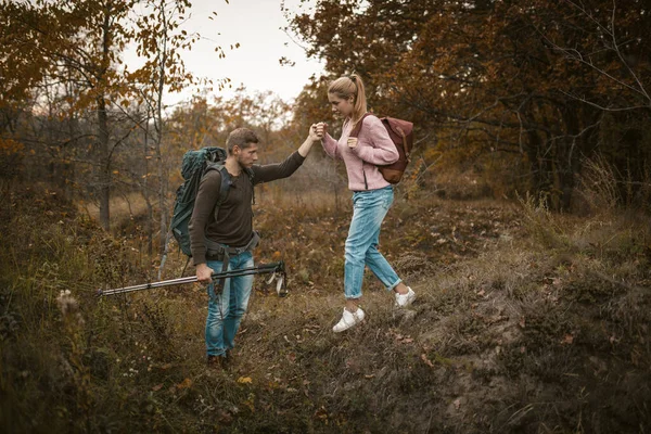 Wandelen in de natuur. Jonge man en vrouw backpackers dalen af van de helling hand in hand en houden wandelstokken in hun handen. Toeristen met rugzakken lopen door het herfstbos. Ondersteuningsconcept — Stockfoto