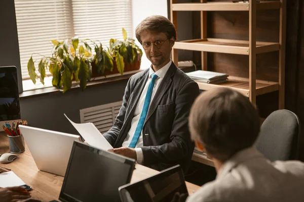 Los colegas masculinos se comunican trabajando en computadoras mientras están sentados en la misma mesa. Reunión de negocios en el acogedor interior de la oficina moderna. Concepto de tiempo de lluvia de ideas — Foto de Stock