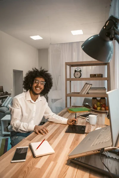 Uomo creativo felice di lavorare da casa. Intelligente ragazzo arabo sorride seduto alla scrivania con un tablet grafico e un computer su di esso in interni accogliente home office. Immagine tonica — Foto Stock