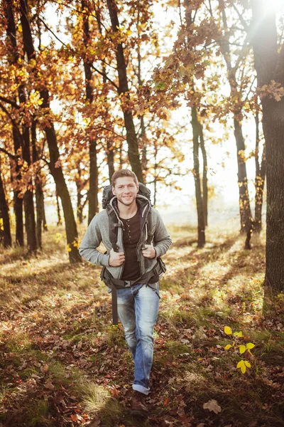 Un joven con una mochila viaja a través del bosque de otoño en un día soleado y cálido. Sonrientes caminatas turísticas caucásicas en la naturaleza. Concepto de senderismo — Foto de Stock