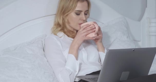 Blondynka pracująca na laptopie pije filiżankę gorącego kakao lub herbaty przed snem po długim ciężkim dniu. Bizneswoman relaksuje się w łóżku po długim locie. Koncepcja biznesu. Prores 422 — Wideo stockowe