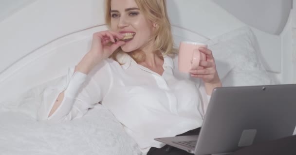 Freelancer werkt aan laptop eten cookie en geniet van een kopje warme chocolademelk of thee tijdens de lange werknacht. Zakenvrouw ontspant in bed na een lange vlucht. Bedrijfsconcept. Prores 422 — Stockvideo