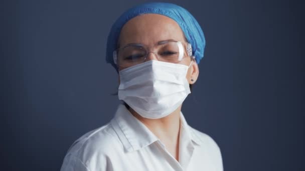 Happy doctor in protective mask standing playfully moving while looking at camera isolated on blue background. Caucasian female medic looks at camera. Copy space on left side. Prores 422 — Stock Video